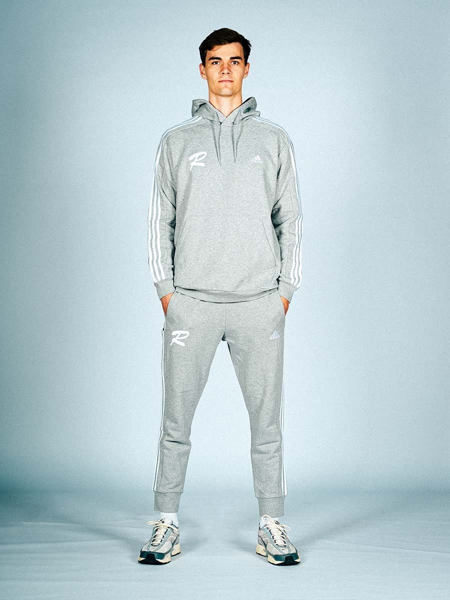 Junger Mann in grauem Adidas-Sportoutfit mit Hoodie, Jogginghose und Turnschuhen, steht auf neutralem Hintergrund, Hände in den Taschen, Blick direkt in die Kamera.
