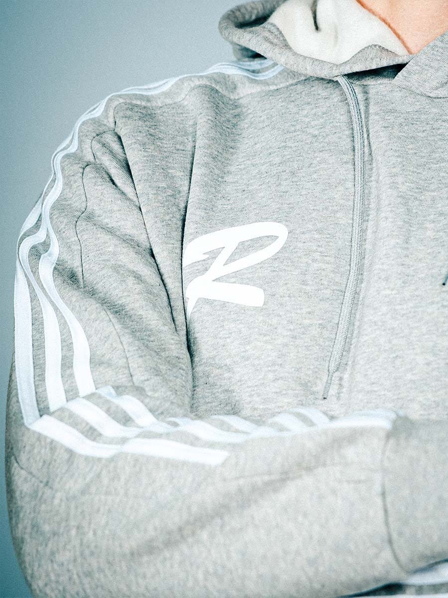 Grauer Kapuzenpullover mit weißen Streifen an den Ärmeln und einem weißen Logo auf der Brust, mit Reißverschluss vorne.