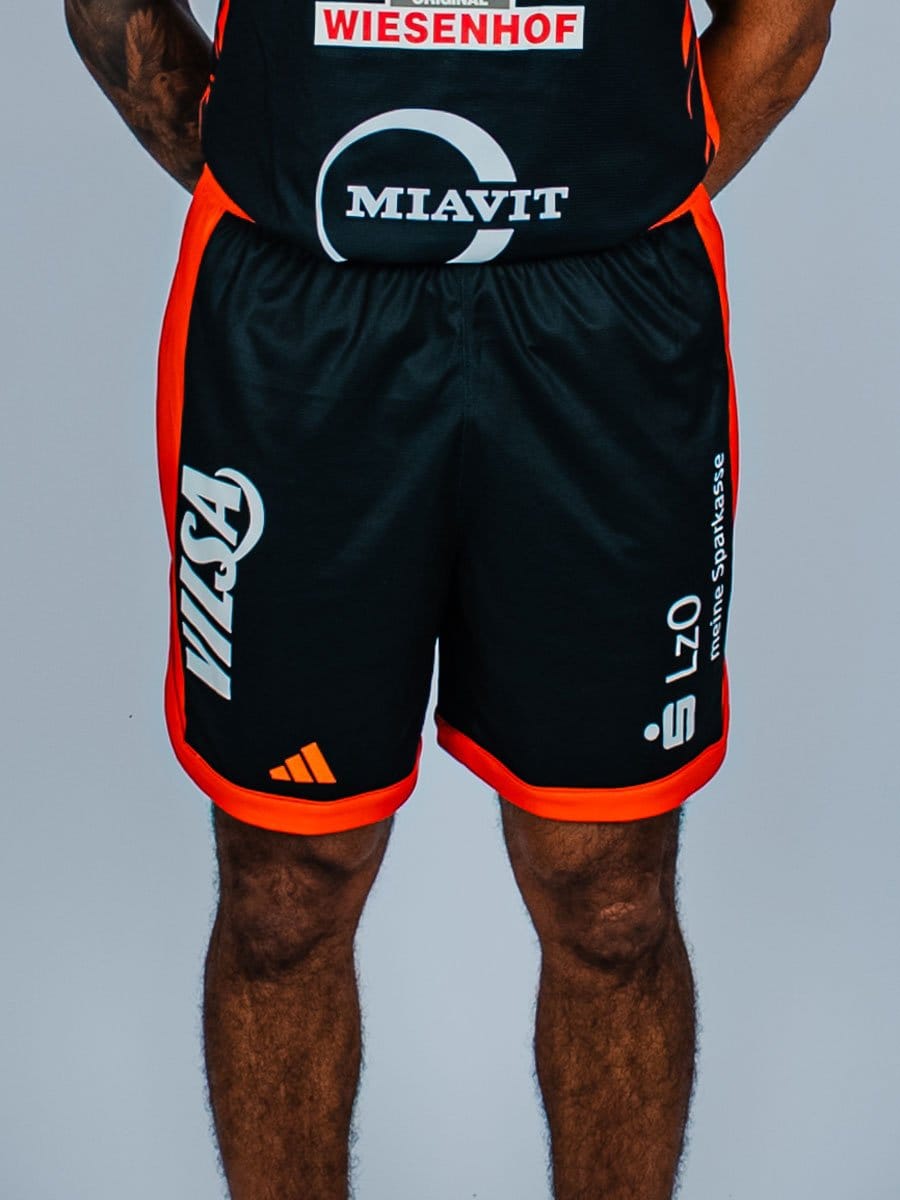 Mann trägt schwarze Sportshorts mit roten Akzenten und weißen Logos, steht vor hellem Hintergrund, nur unterhalb der Taille sichtbar.