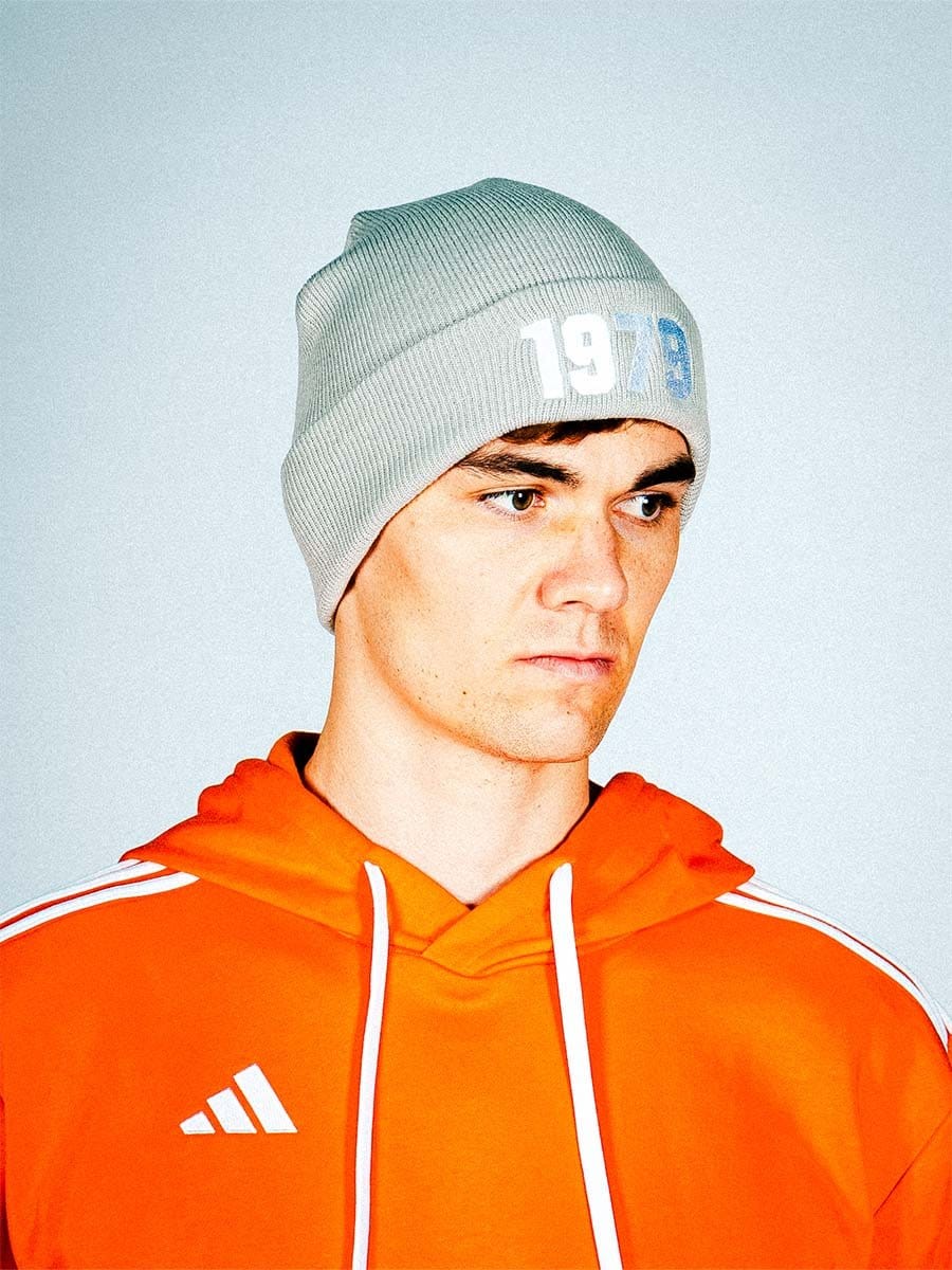 Junge Person trägt grauen Strickmütze mit weiß-blauer Aufschrift und orange Adidas-Hoodie, blickt nach rechts, neutraler Gesichtsausdruck, heller Hintergrund.