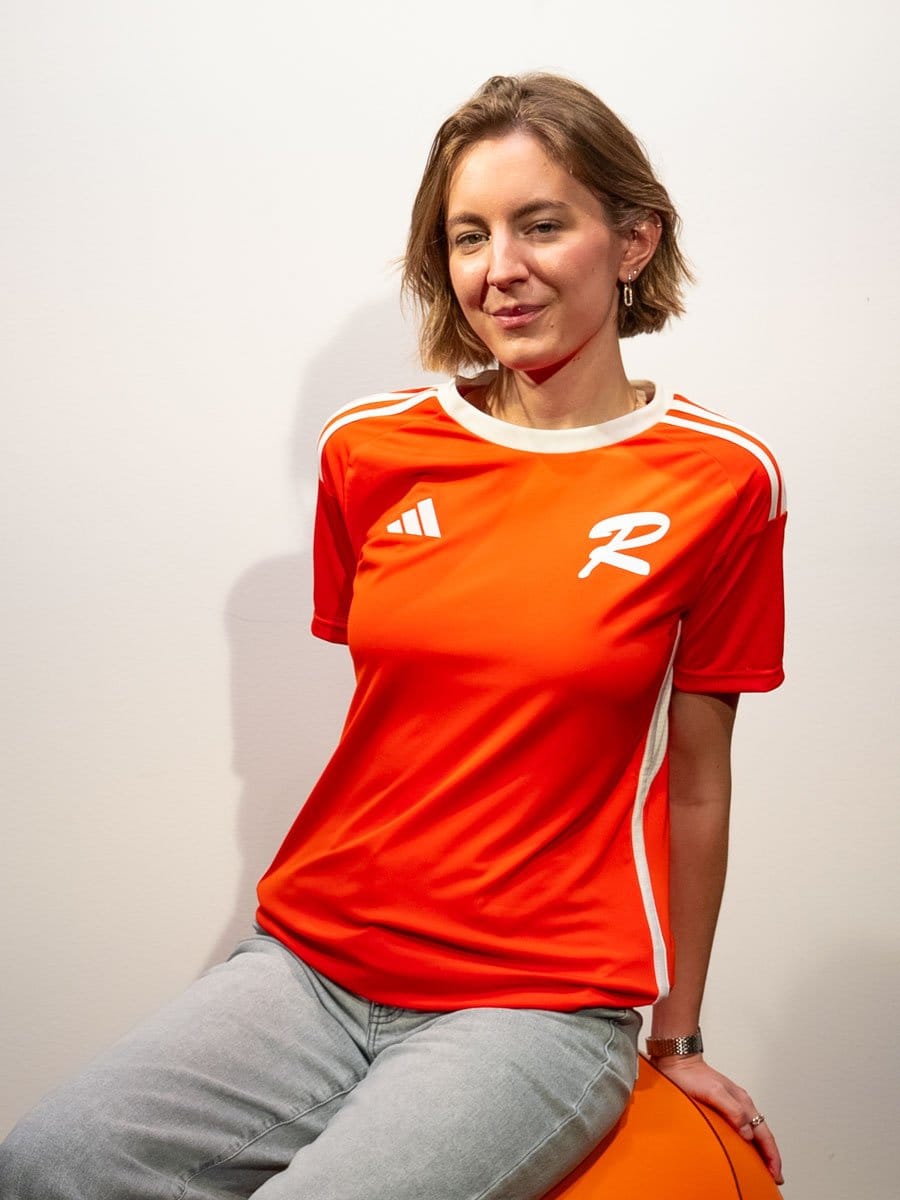 Junge Frau mit kurzen braunen Haaren sitzt auf einem orangefarbenen Ball, trägt ein rotes Sportshirt mit weißen Streifen und graue Jeans vor einer weißen Wand.