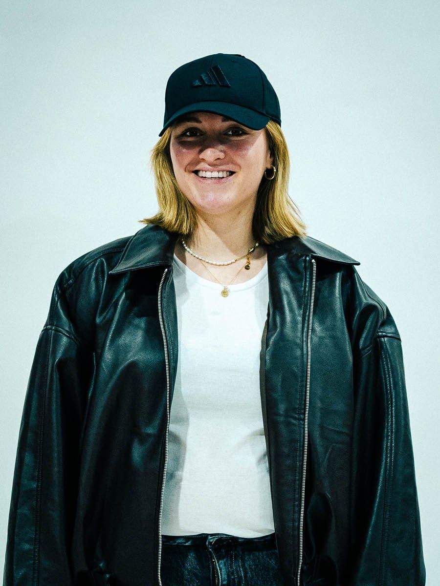Junge Frau trägt schwarze Adidas-Cap, schwarze Lederjacke, weiße T-Shirt, goldene Halsketten, kleine Ohrringe und lächelt vor hellem Hintergrund.