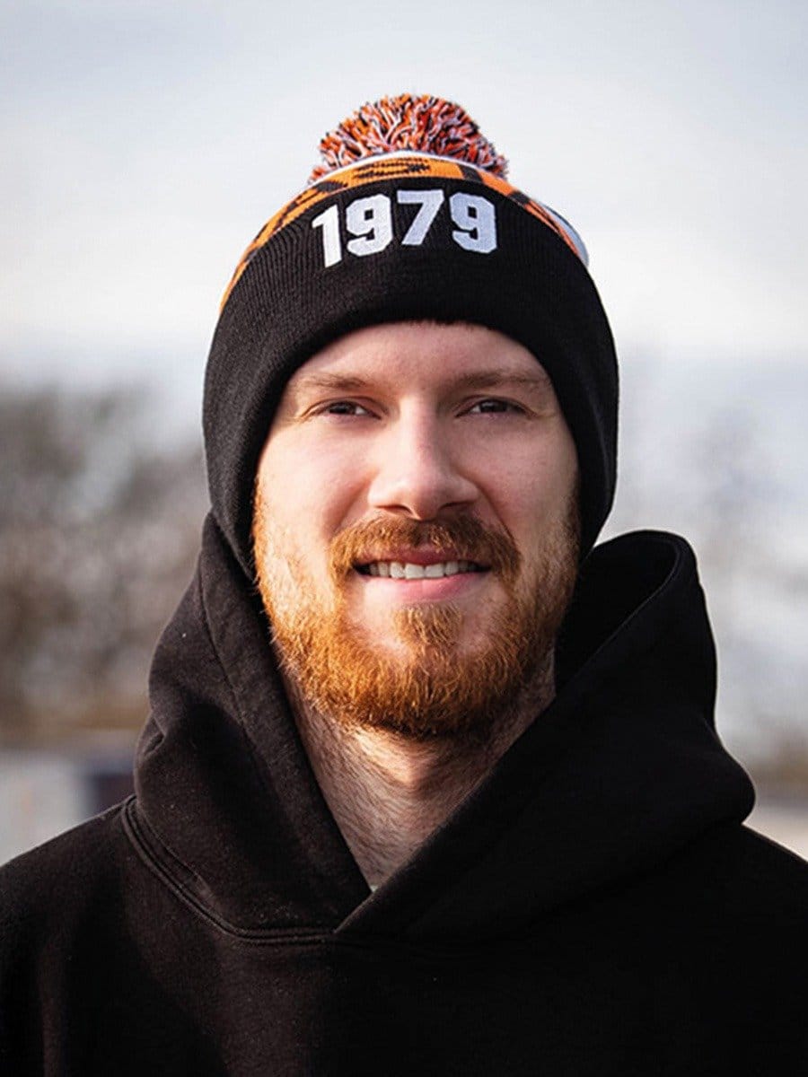 Junge Mann mit rotem Bart trägt schwarze Kapuzenjacke und eine schwarze Mütze mit "1979" und buntem Bommel, steht draußen bei bewölktem Himmel.