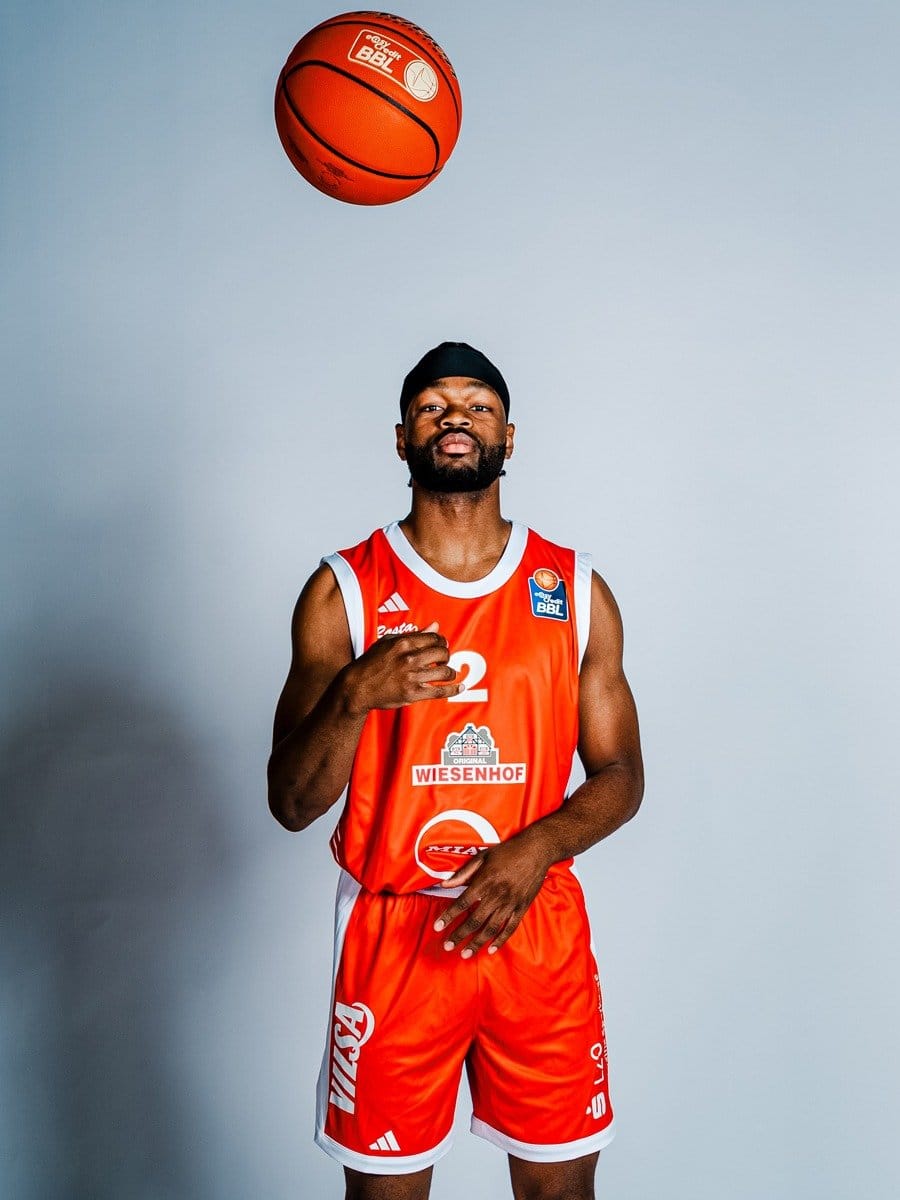 Basketballspieler in rotem Trikot und Shorts, trägt eine schwarze Mütze, steht vor hellem Hintergrund, hält eine Basketball im Blick, der in der Luft schwebt.