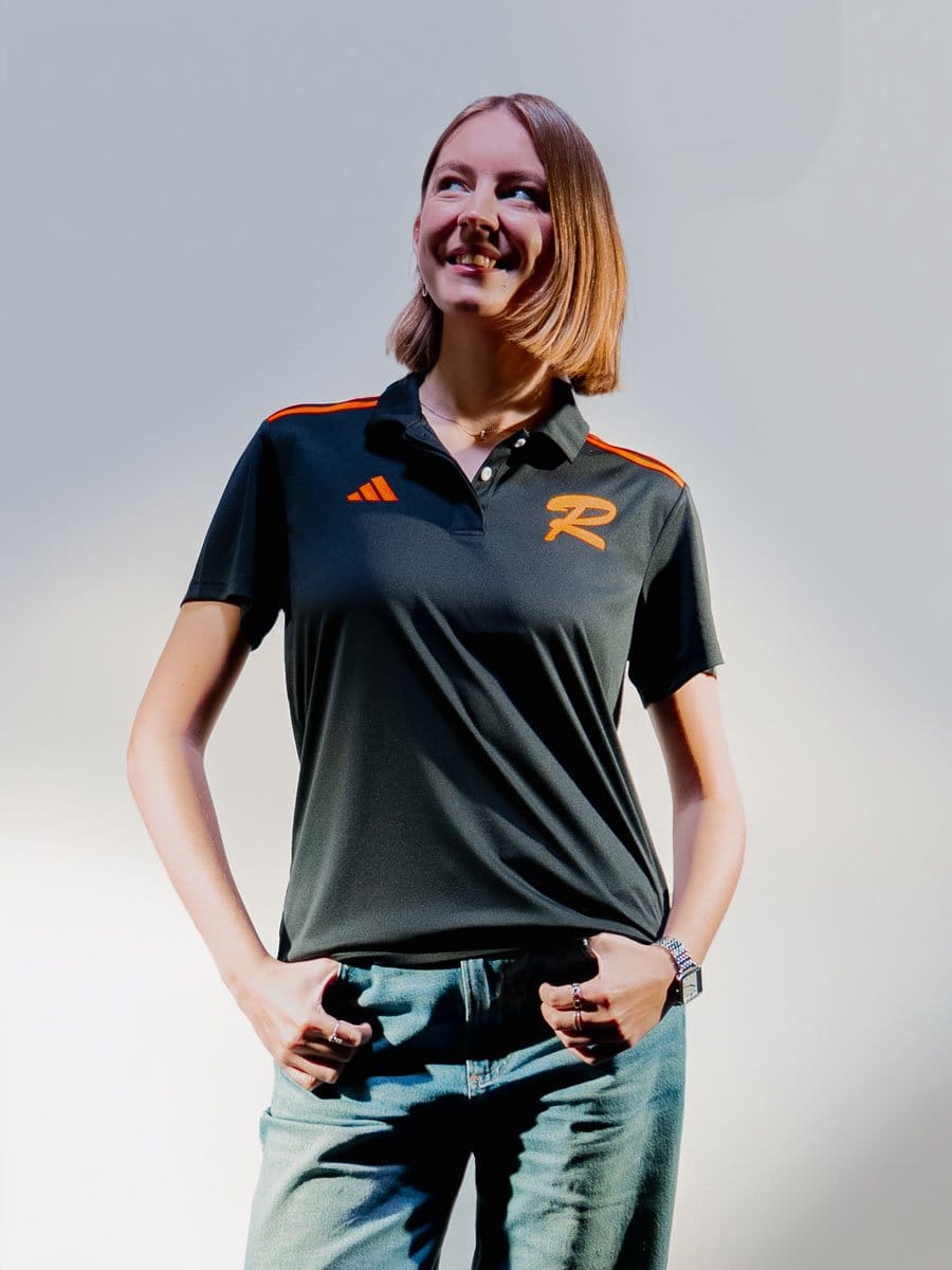 Junge Frau mit Schulterlänge brauner Haar, trägt schwarze Polo mit orangefarbenen Akzenten und einem "R" Logo, lächelt, steht vor hellem Hintergrund.