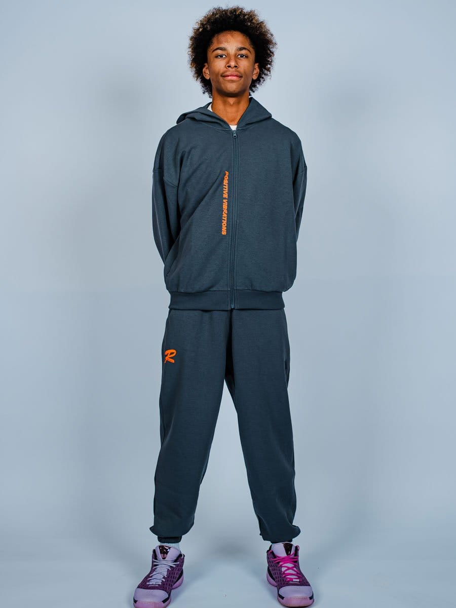 Junge Person in dunklem Sportoutfit mit Hoodie und Jogginghose, trägt pink-lila Sportschuhe, steht vor hellem Hintergrund, Blick nach vorne, lässige Haltung.