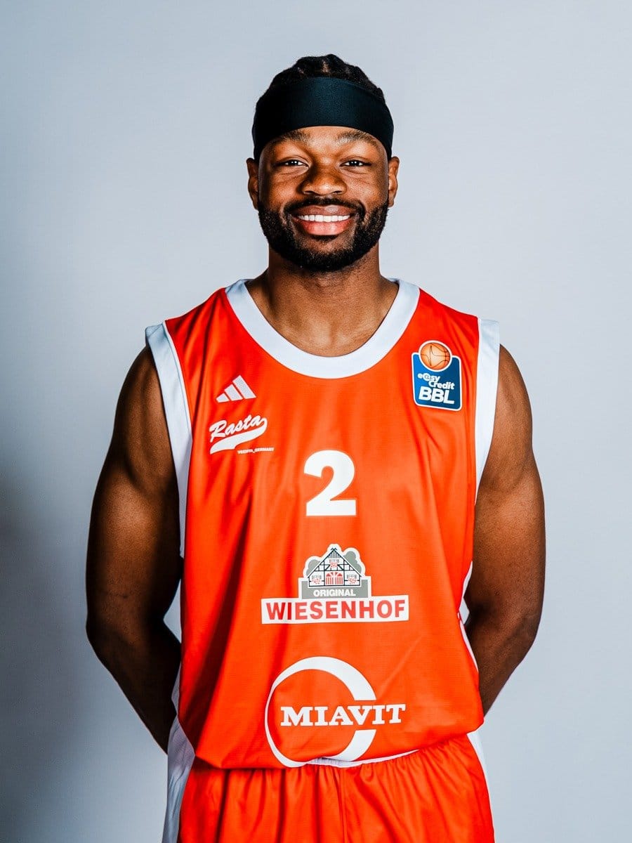 Ein lachender Basketballspieler in rotem Trikot mit schwarzen Haaren, dunklem Bart, schwarzen Stirnband, vor hellem Hintergrund.