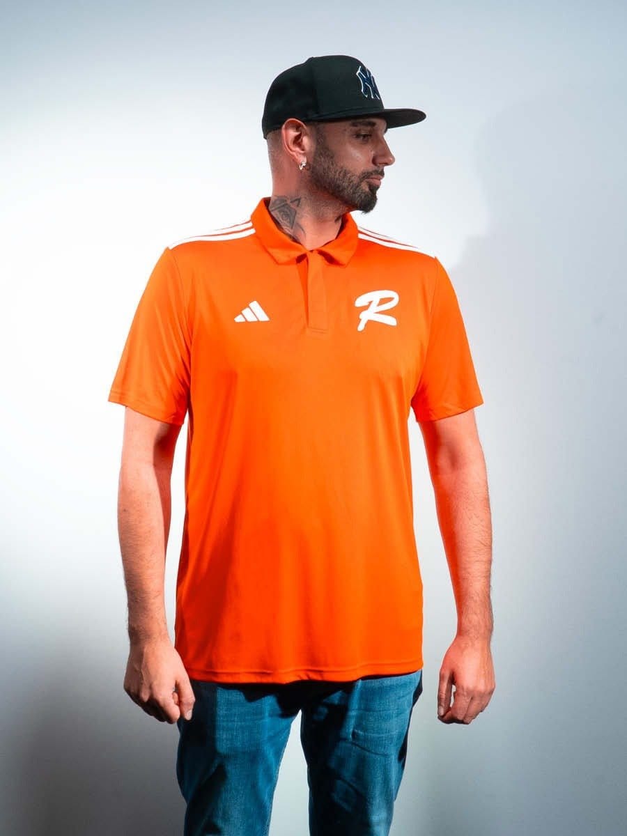 Mann trägt orange Sporttrikot mit weißen Logos, schwarze Cap, blaue Jeans, steht seitlich vor hellem Hintergrund.