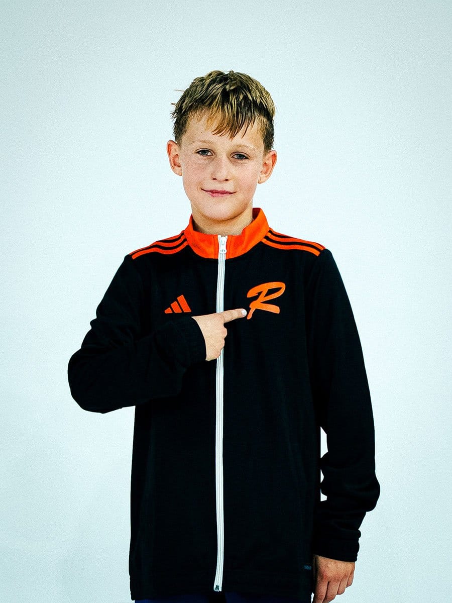 Junge trägt schwarze Sportjacke mit orangeem Reißverschluss, orange-gestreiftem Schulterdesign und einem orangefarbenen "R" Logo, zeigt auf das Logo.