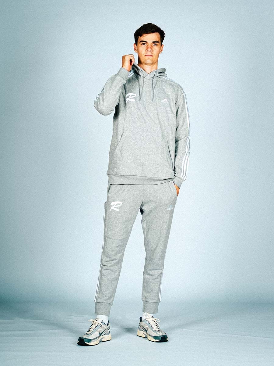 Junger Mann in grauem Adidas-Sportoutfit mit Hoodie, Jogginghose und Sneakers, steht vor hellem Hintergrund, schaut ernst in die Kamera, rechte Hand an der Schulter.