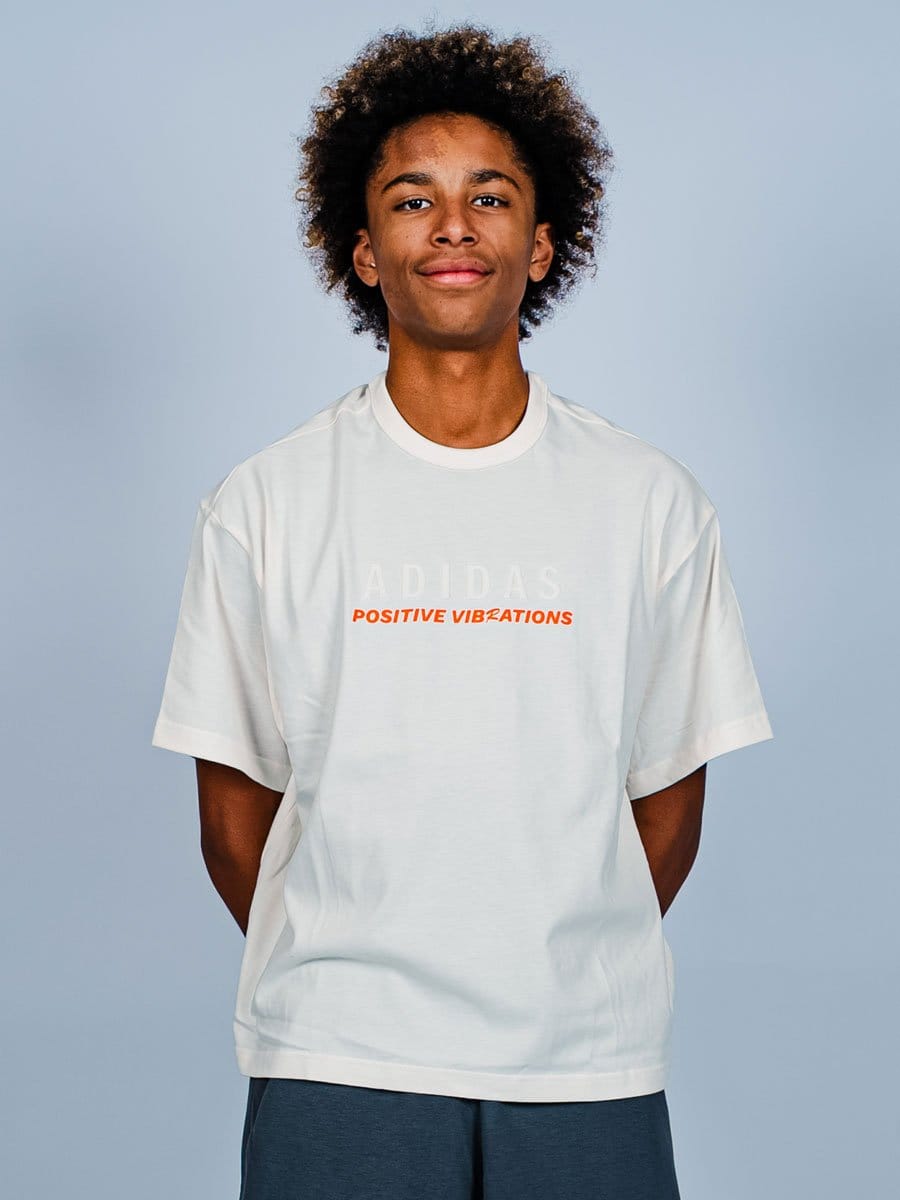 Junge Person mit lockigem Haar trägt weiße T-Shirt mit "ADIDAS" und "POSITIVE VIBRATIONS" in orange, steht vor hellem grauem Hintergrund, lächelt leicht.