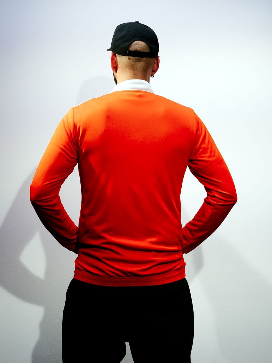 Mann trägt eine rote Sportjacke, schwarze Hose und eine schwarze Cap, steht mit dem Rücken zur Kamera vor einer weißen Wand.