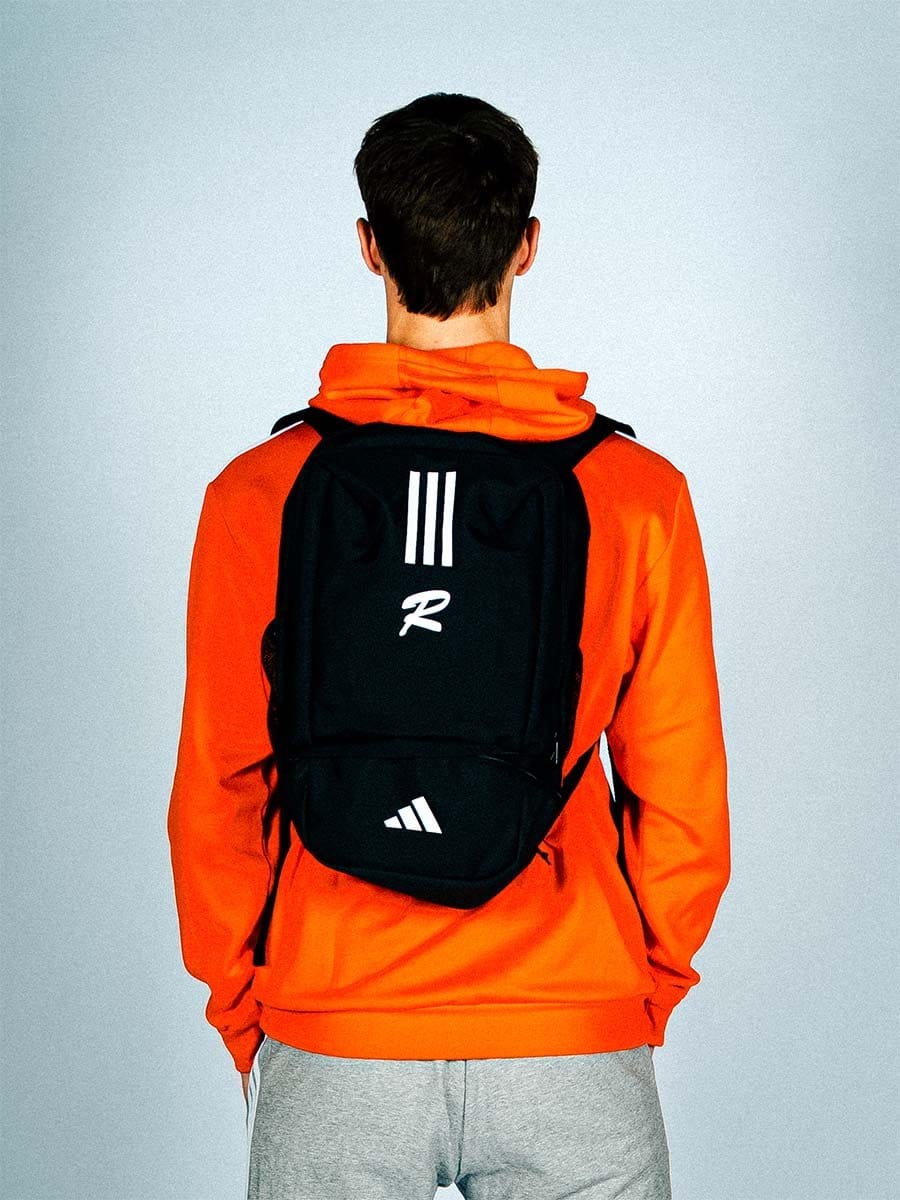 Junge Person von hinten trägt eine schwarze Adidas-Schultasche und einen orangefarbenen Hoodie vor einem hellgrauen Hintergrund.