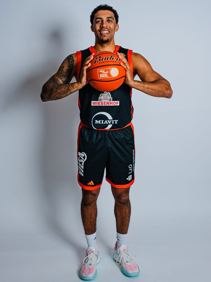 Basketballspieler in schwarzem Trikot mit roten Akzenten, hält einen Basketball vor der Brust, trägt weiße Socken und bunte Turnschuhe, steht vor grauem Hintergrund.