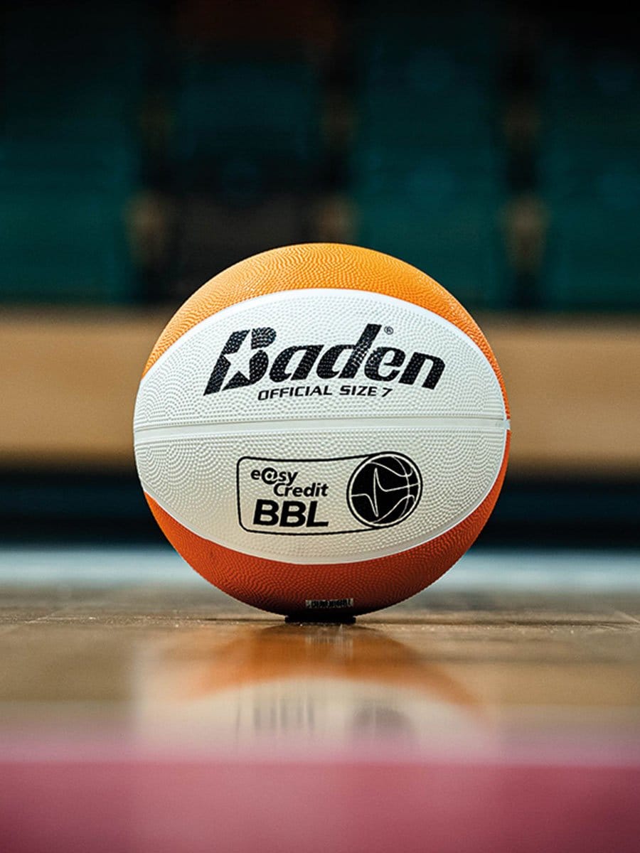 Basketball mit orange-weißer Oberfläche und schwarzen Schriftzügen liegt auf einem Basketballfeld, im Hintergrund sind leere blaue Sitzreihen sichtbar.