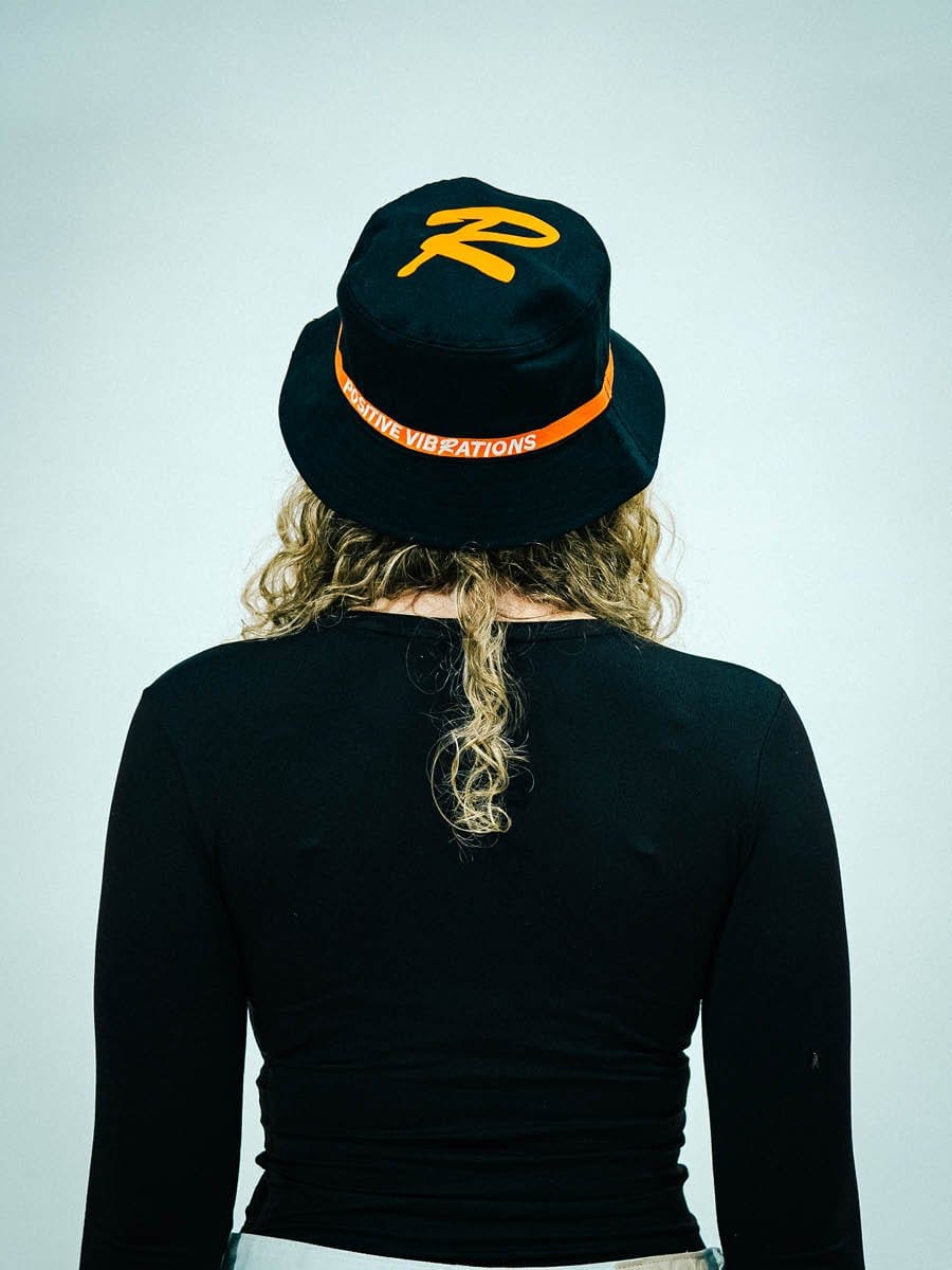 Person mit lockigem Haar trägt schwarzen Hut mit gelbem Logo und orangefarbenem Band mit Schriftzug "POSITIVE VIBRATIONS" auf schwarzem Oberteil.