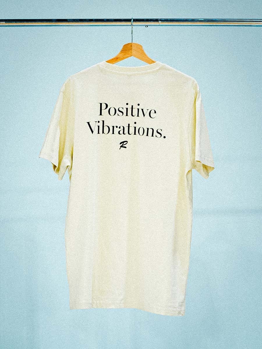 Weißes T-Shirt mit schwarzem Text "Positive Vibrations." und einem kleinen Logo, hängt an einem Holzkleiderbügel an einer Stange vor einem hellblauen Hintergrund.