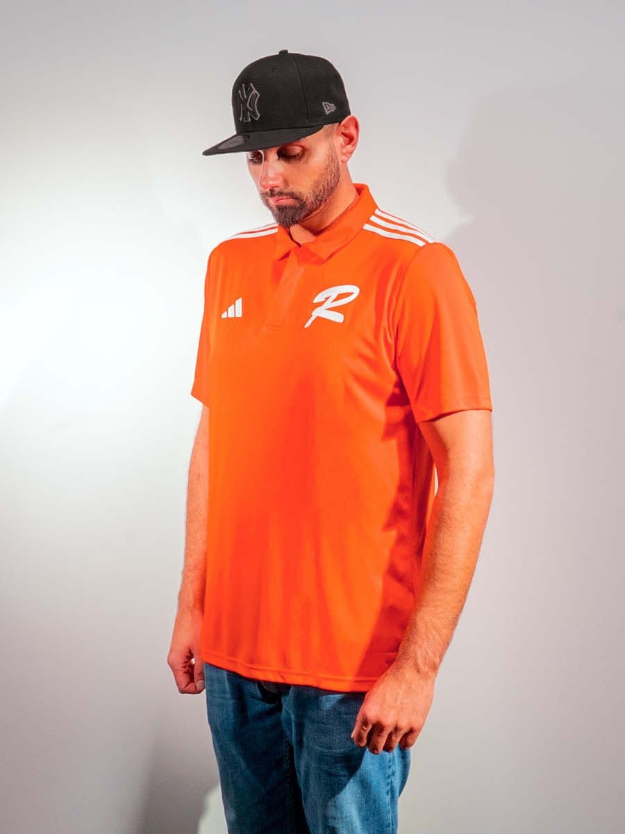 Mann trägt eine schwarze Baseballcap, ein orangefarbenes Sportshirt mit weißen Logos und blaue Jeans, steht vor hellem Hintergrund.