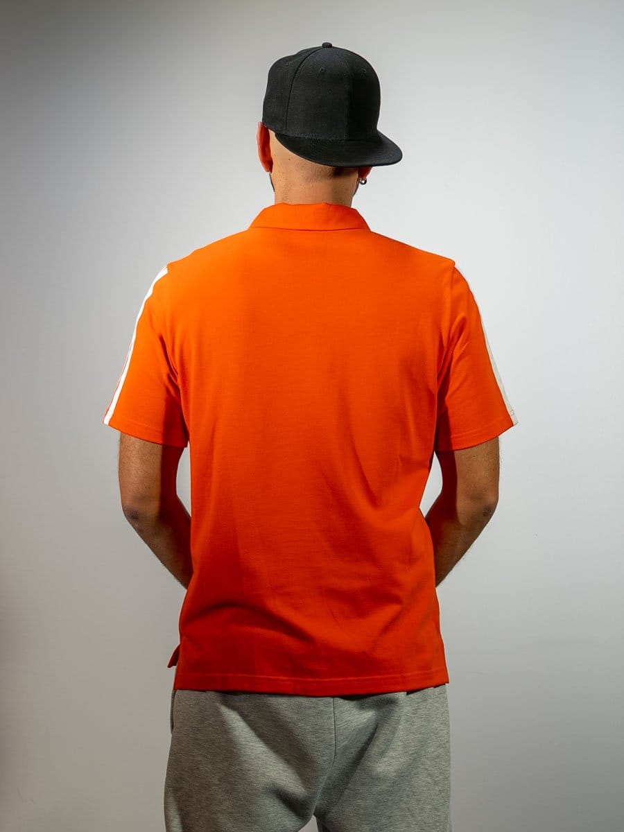 Mann trägt eine schwarze Cap, eine orange Sportjacke mit weißen Streifen an den Ärmeln und graue Jogginghose, steht vor weißer Wand, von hinten fotografiert.