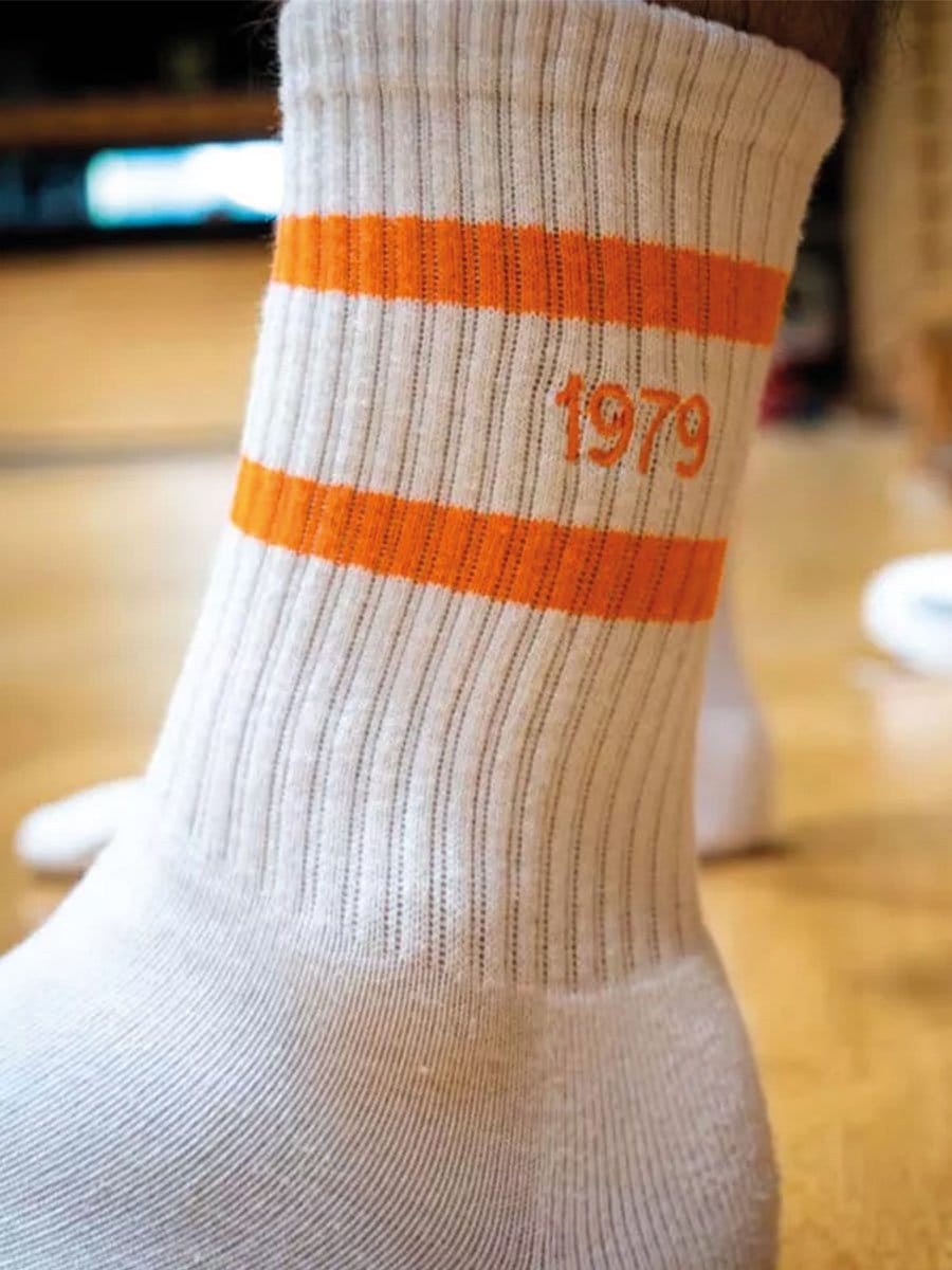 Socken in beige mit orange-weißen Streifen und der Jahreszahl 1979 in orange, getragen am Fuß eines Menschen, im Hintergrund ein Wohnzimmer.