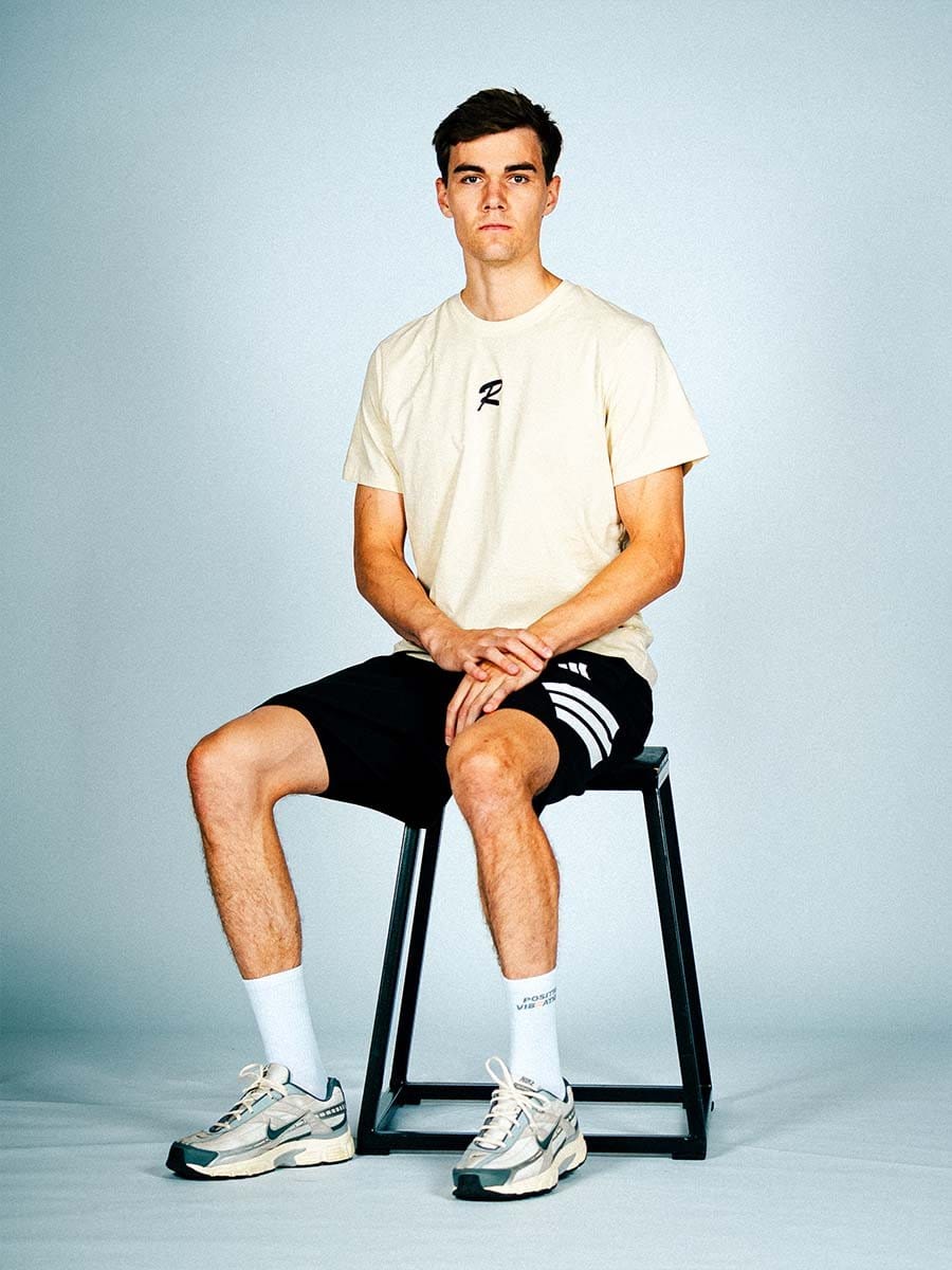 Junger Mann sitzt auf einem schwarzen Hocker, trägt ein cremefarbenes T-Shirt, schwarze Shorts, weiße Socken und Sneakers, vor hellem Hintergrund.