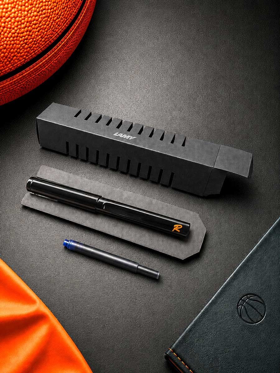 Schwarzer Filzstift, schwarzer Kunststoff-Box mit Ventilationsschlitzen, orange-weißer Schriftzug, auf schwarzer Tischfläche neben einem Basketball und einem schwarzen Notizbuch.