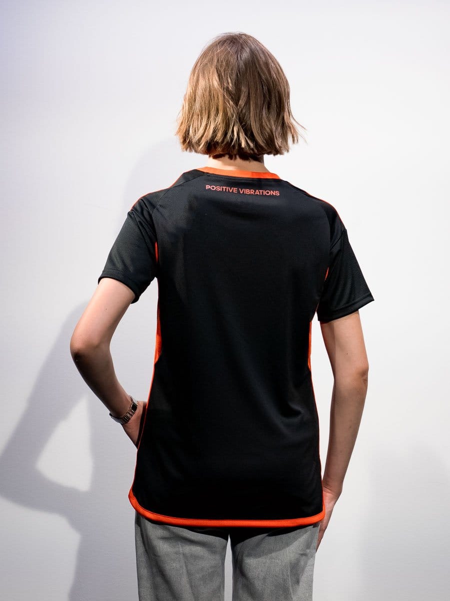 Rückansicht einer Person mit kurzen, braunen Haaren, die ein schwarzes Sportshirt mit roten Akzenten trägt, auf dem "Positive Vibrations" steht.