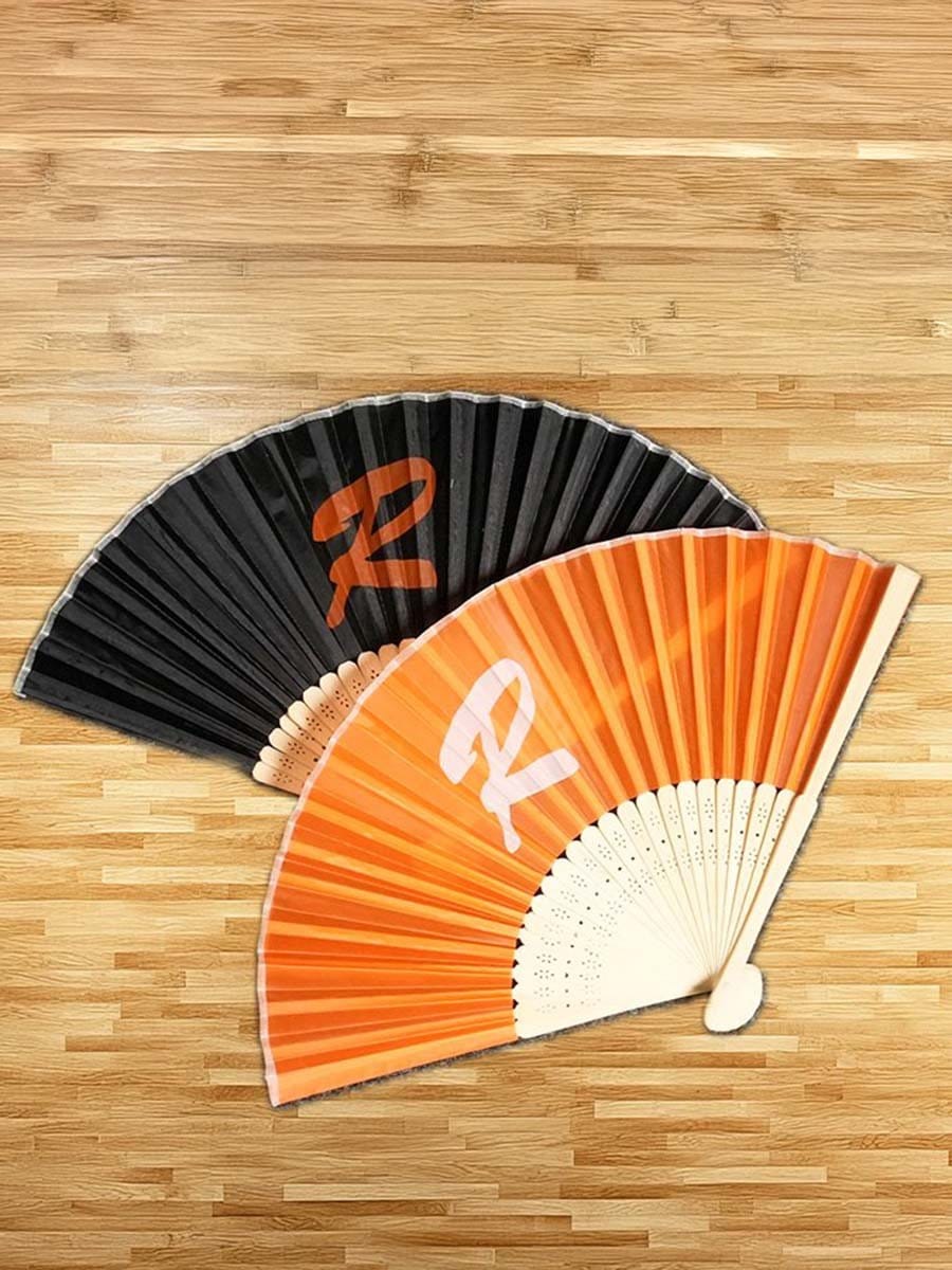 Zwei handgehaltene Fächer, einer schwarz mit orangeem "R" und einer orange mit weißem "R", liegen auf einem Holztisch.