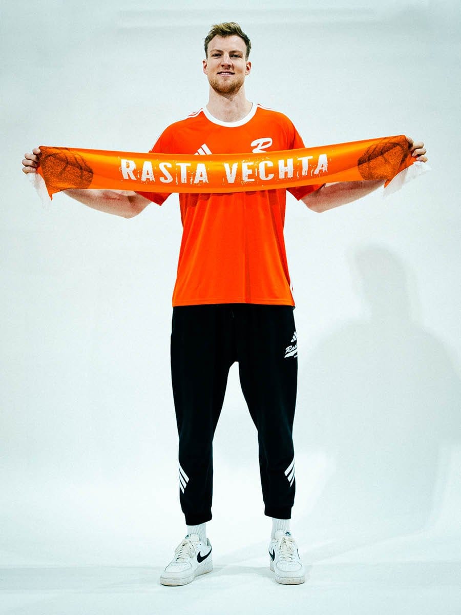 Junger Mann in rotem Sportshirt und schwarzen Jogginghosen hält einen orangefarbenen Schal mit weißen Buchstaben "RASTA VECHTA" vor der Brust, vor weißem Hintergrund.