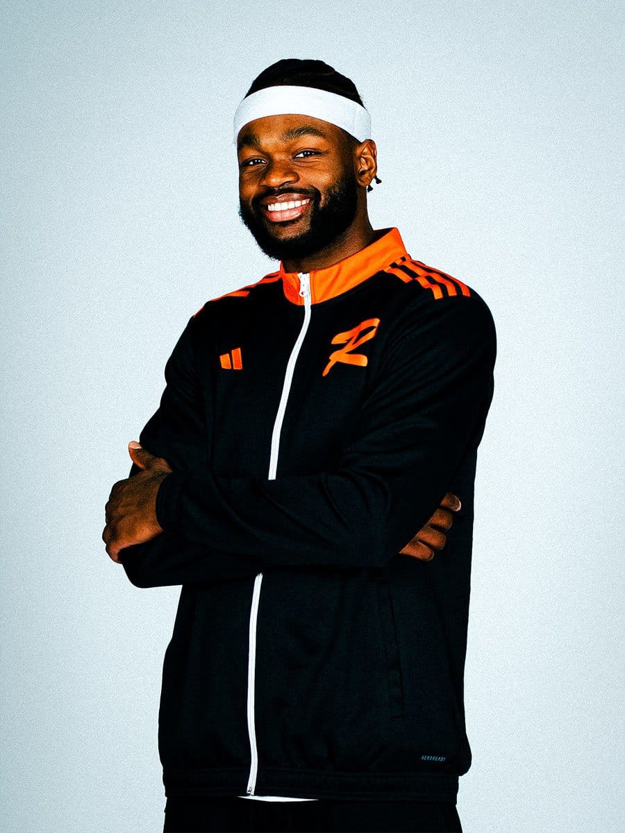 Junger Mann mit dunklem Haar, Bart und weißen Stirnband, trägt schwarze Sportjacke mit orange Akzenten, lächelt, Arme verschränkt vor weißem Hintergrund.