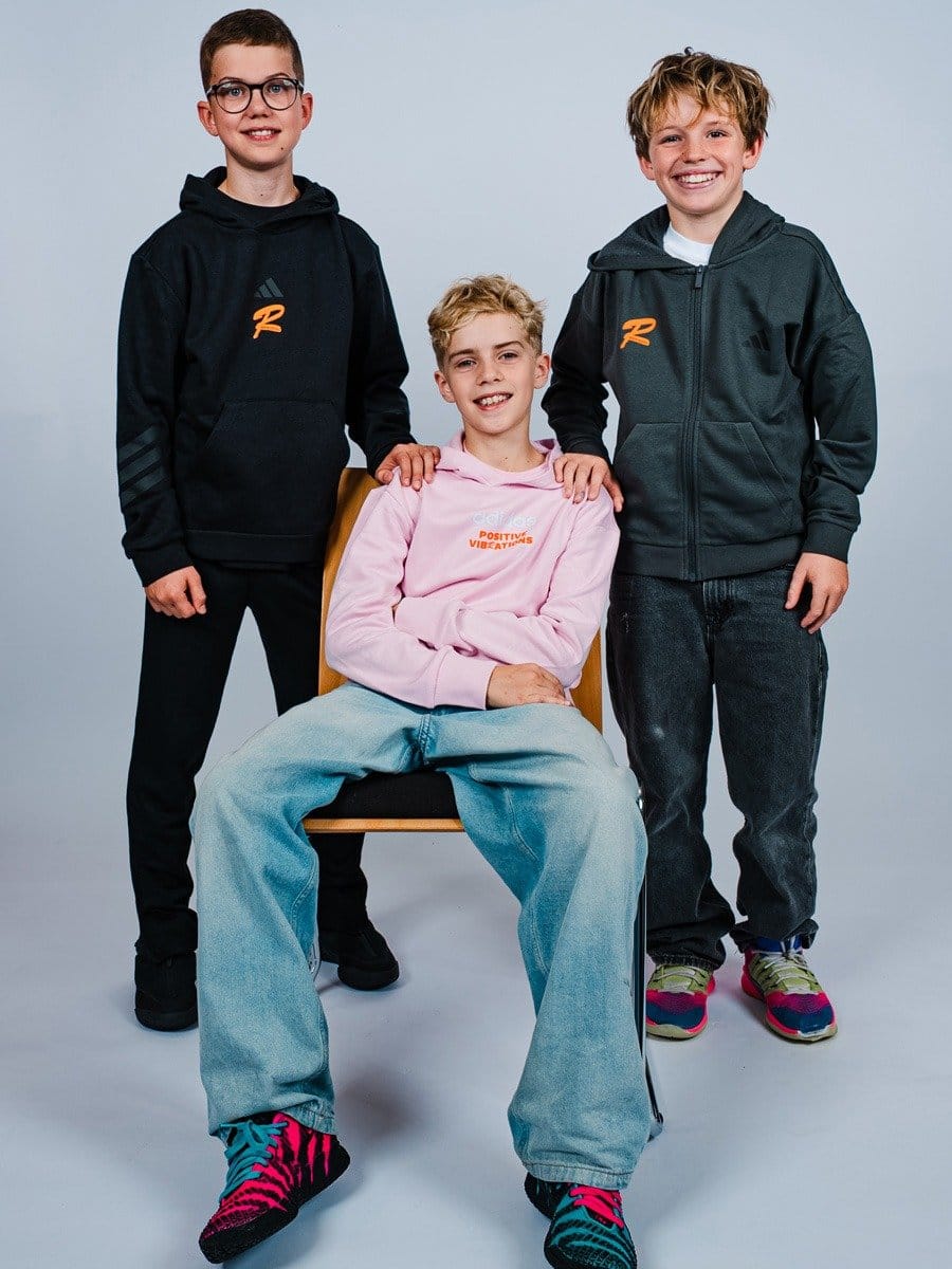 Drei lachende Jungen, einer sitzt auf einem Stuhl, die anderen stehen hinter ihm. Alle tragen sportliche Hoodies und bequeme Hosen vor einem neutralen Hintergrund.
