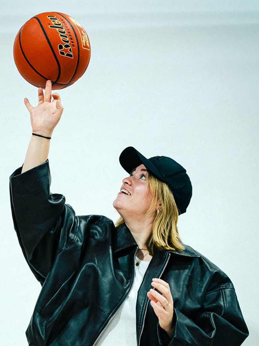Junge Frau in schwarzer Lederjacke und Cap wirft einen Basketball nach oben, blickt dabei nach oben, vor einem hellen Hintergrund.