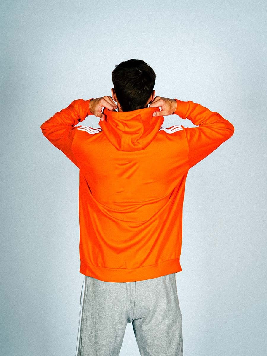 Junger Mann von hinten, trägt orange Kapuzenjacke und graue Jogginghose, steht vor hellem Hintergrund, hält die Kapuze mit beiden Händen.