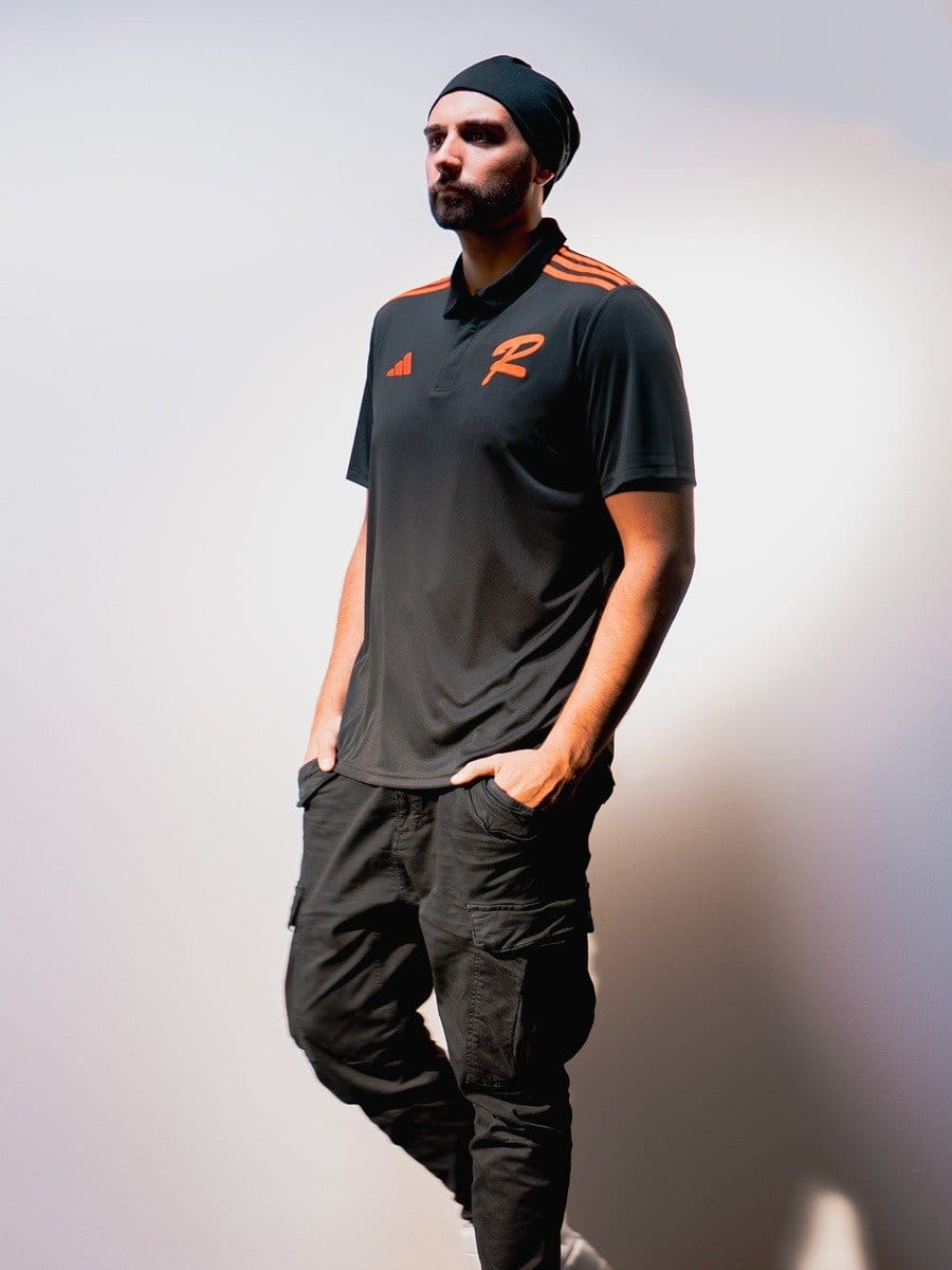 Mann in schwarzem Sportoutfit mit orangefarbenen Akzenten, trägt eine schwarze Mütze, steht mit Händen in den Taschen vor weißem Hintergrund.
